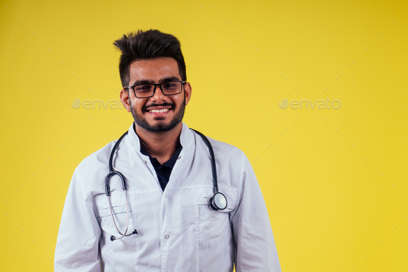 indian male doctor wearing glasses and white cloak in yellow background studio