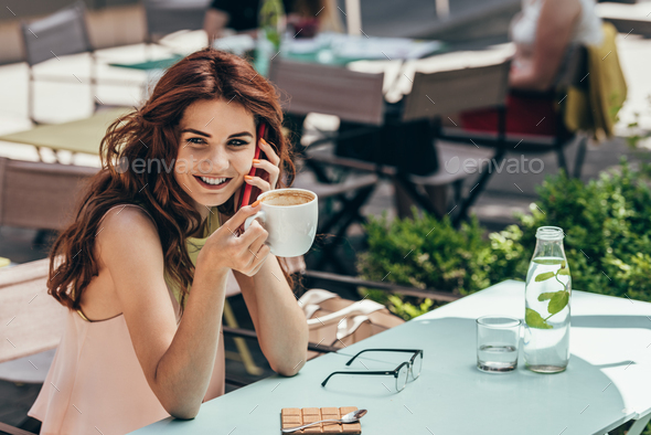 portrait of smiling woman with cup of coffee talking on smartphone in cafe