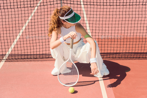 fashionable woman in white clothing and cap with tennis racket sitting at tennis net on court