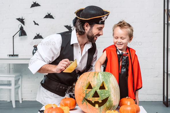 happy man in pirate costume and son in vampire halloween costume with pumpkin together at home