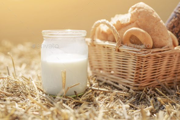A jar of fresh milk in a field on straw next to a basket filled with bread products.