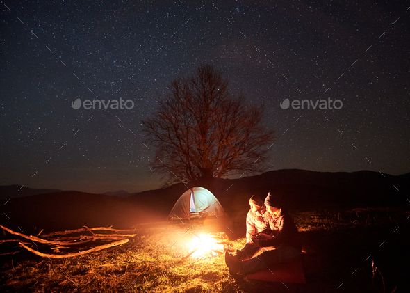 Night camping in mountains. Couple hikers having rest near campfire, tourist tent under starry sky