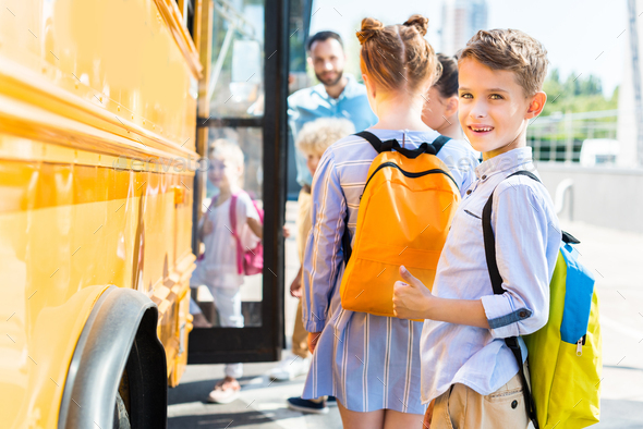 smiling little schoolboy entering school bus with classmates while teacher standing near door