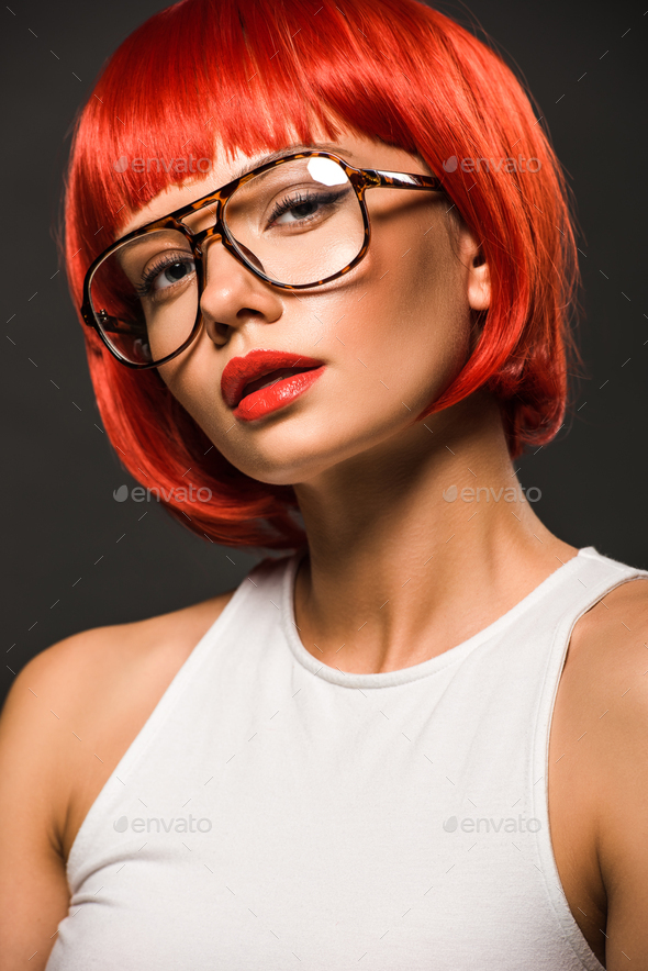 attractive young woman with red bob cut and stylish eyeglasses looking at camera isolated on grey