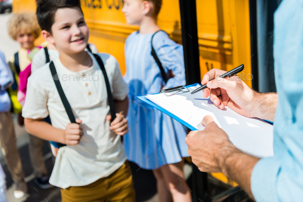 cropped shot of teacher writing in list on clipboard while pupils entering at school bus blurred on