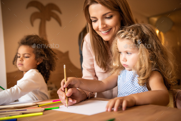 happy tutor helping kid drawing in kindergarten