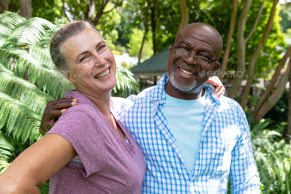 Portrait of happy multiracial senior couple standing with arms around shoulders in backyard