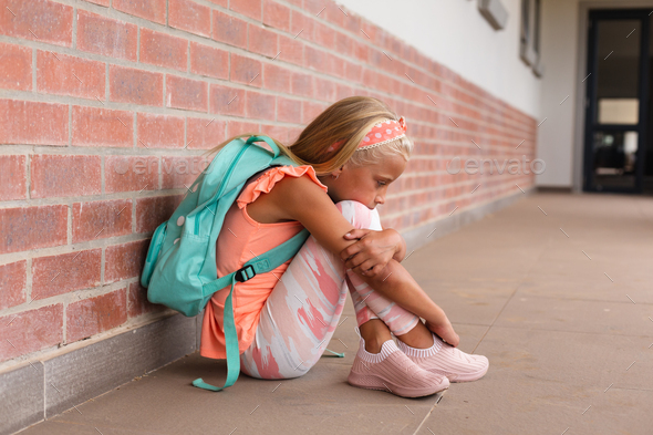 Full length of sad caucasian elementary schoolgirl hugging knees while sitting on floor in school