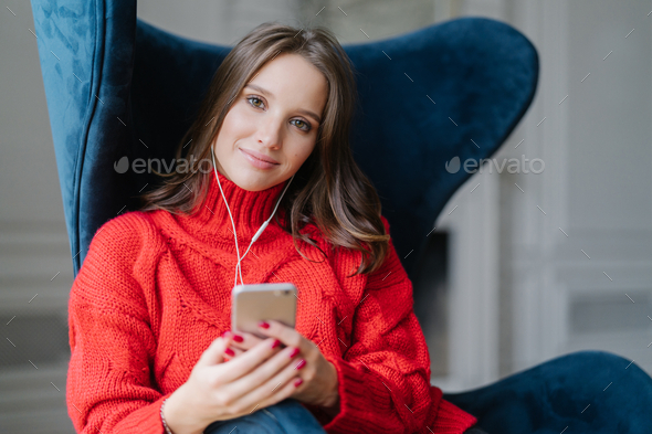 Satisfied female melamine enjoys favourite music, listens audio record, dressed in red sweater.