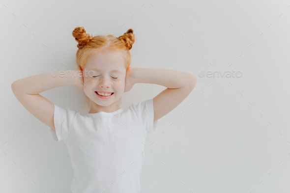 Positive good looking ginger freckled beautiful girl covers ears with hands, hears very loud music.