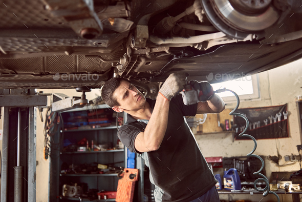 Repair man fixing car in repair station under lifted car during repair in garage