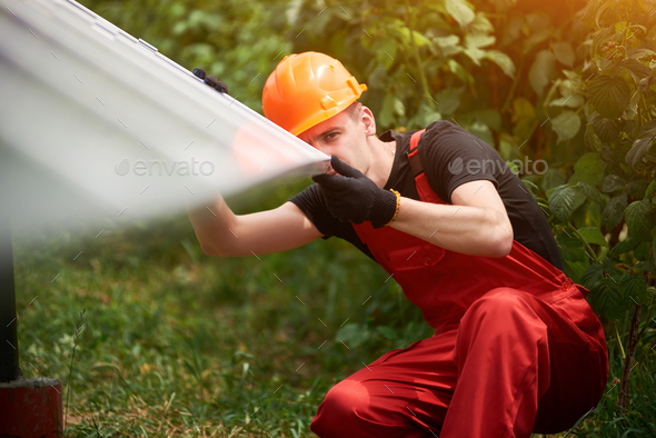 Worker in orange uniform, safety cap near on blurry background of solar panels and greenery.