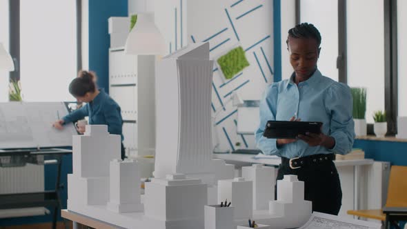 African American Engineer Holding Tablet to Work on Building Model and Maquette alt
