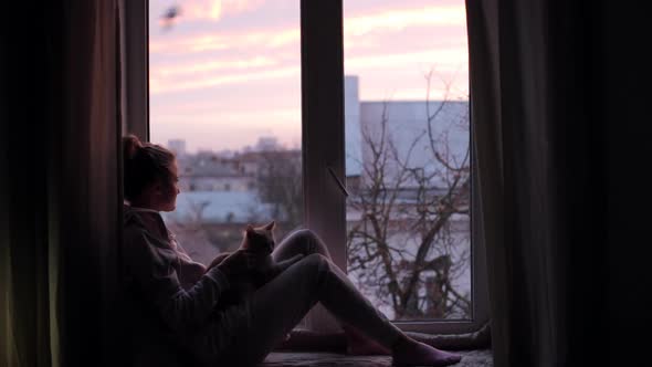 Girl Watches the Pink Dawn Sitting on the Windowsill with Her Cat