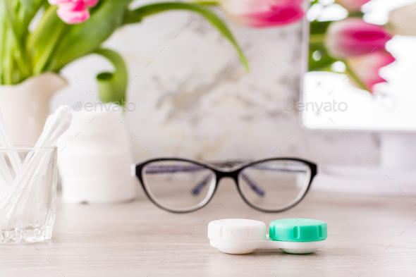 Container for lenses on the background of glasses, hygiene products and a mirror on the table
