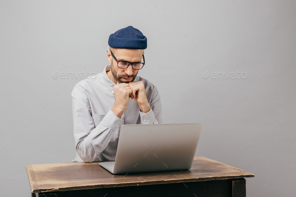 Serious male workaholic sits at desktop with modern electronic device, watches video.