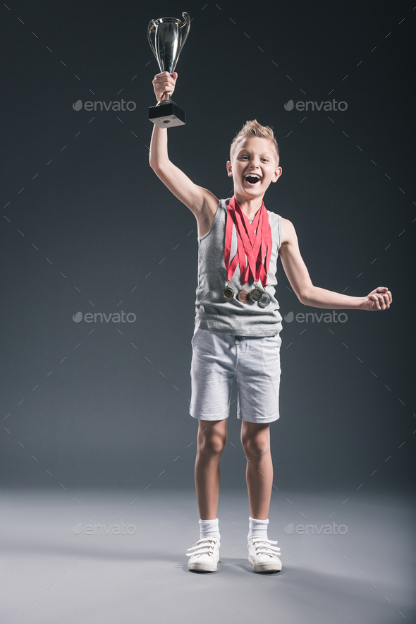 happy boy in sportswear with medals and champions cup gesturing on dark background