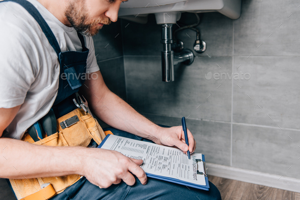 cropped shot of male plumber with toolbelt writing in clipboard near broken sink in bathroom