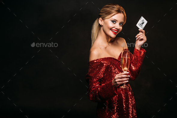 happy attractive girl in red shiny dress holding joker card and glass of champagne isolated on
