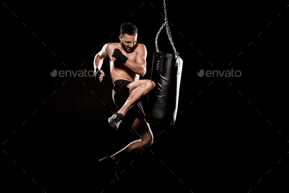 shortless man performing flying kick near punching bag isolated on black