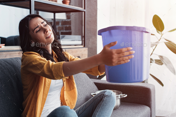 Upset woman holding bucket under leaking ceiling in living room