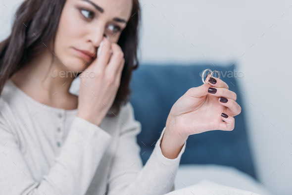 sad woman holding wedding rings and wiping tears while crying at home