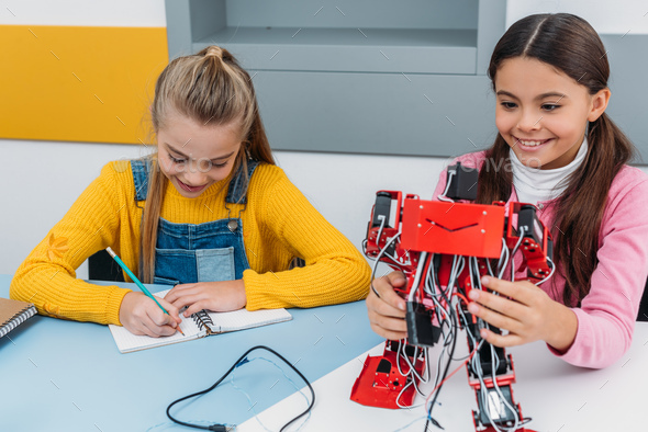 smiling schoolgirls writing in notebook and holding red robot at desk in stem education class