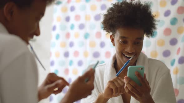 Afro Man in Bathrobe Brushing Teeth and Using Mobile Phone in Bathroom alt