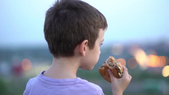 The Boy Eats a Big Juicy Burger and Looks at the Lights of the Evening City