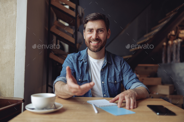 Cheerful attractive man smiling and reaching for camera with hand. Taking gesture. Coffee in cafe.