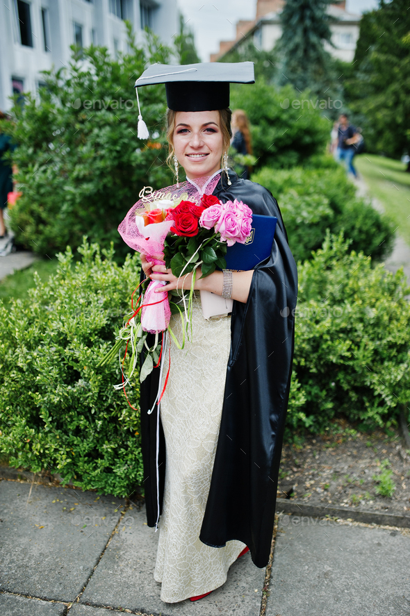 Portrait of a beautiful female graduate in dress and graduation gown with hat holding flowers.