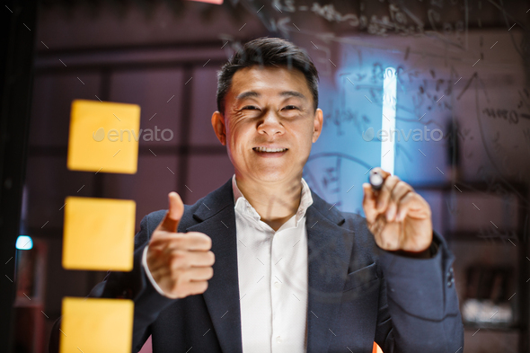 Asian businessman drawing pie chart on glass board with orange sticky notes