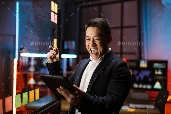 Asian businessman, standing near glass board, looking at the tablet pc screen and celebrating win