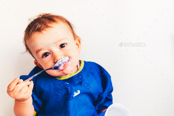 Baby savoring his first yogurt with a spoon, with a spotted face and adorable expression.