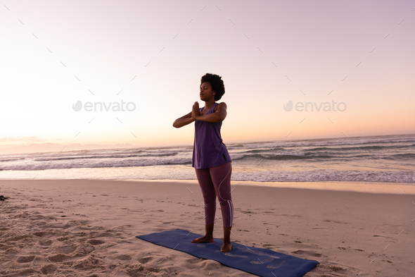 Full length of african american mature woman meditating in prayer pose on mat at beach at sunset