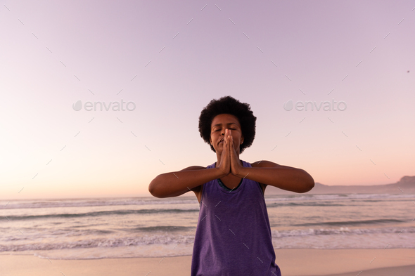 African american mature woman with afro hair meditating in prayer pose at beach against clear sky