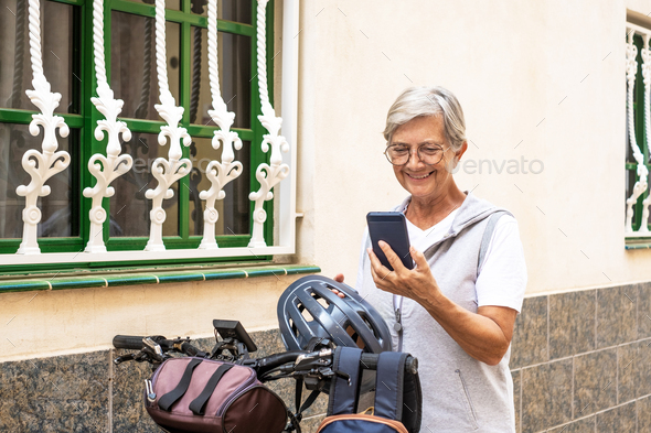 Cyclist elderly woman in urban street holding helmet before running with electro bicycle using phone