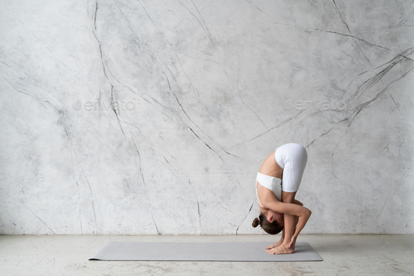Young woman in white outfit practicing yoga standing forward bend, uttanasana