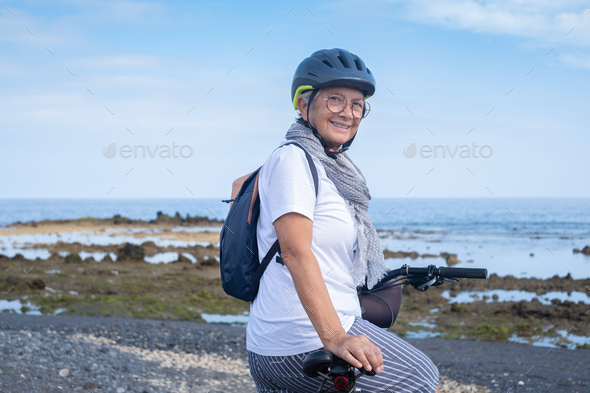 Happy cyclist senior woman wearing helmet and backpack standing along the sea beach