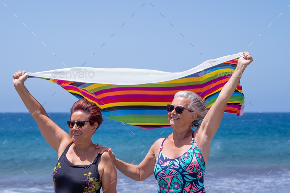 Two mature women enjoying vacation at the sea beach holding rainbow colored towel in the wind