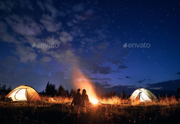 Young couple sitting near campfire under cloudy night sky.