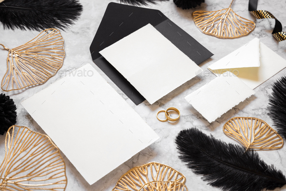 Cards and envelope on marble table near black feathers and golden leaves. Wedding mockup