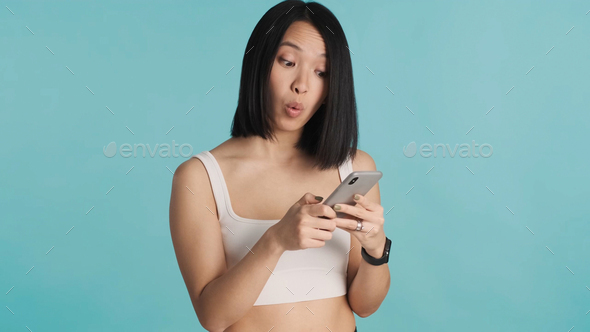 Surprised Asian woman staring at smartphone reading news over blue background. Wow face