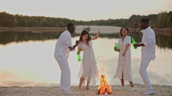 Group of Friends Having Fun and Dancing on a Lake Beach Drinking Near Campfire
