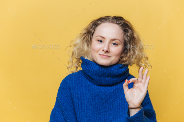 Female with blonde curly hair, satisfied expression, shows ok sign, demonstrates approval.