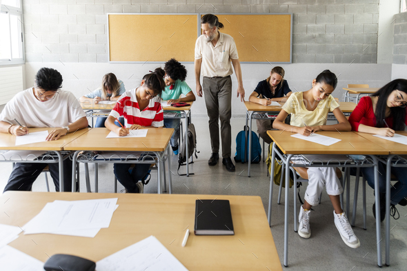 Mature caucasian teacher walking in class to monitor multiracial group of students taking an exam. - Stock Photo - Images