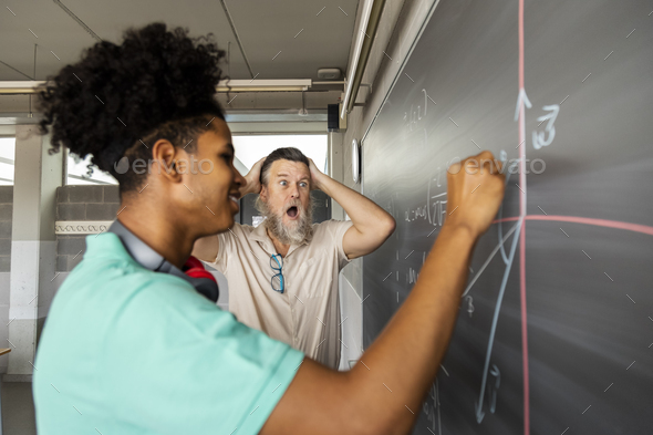 Surprised male teacher with how well teen high school student solves math exercise on blackboard.