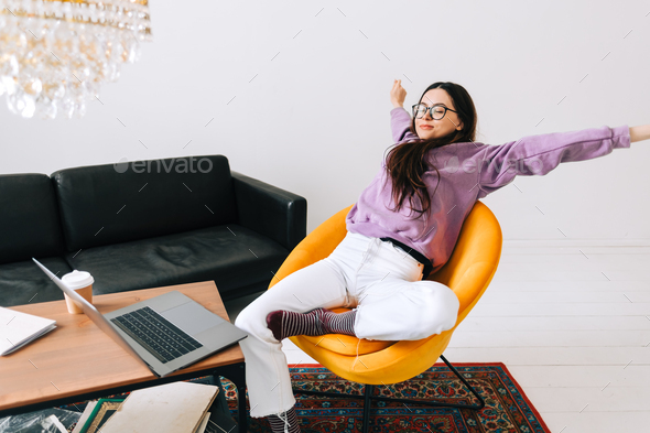 Young caucasian woman relaxing on armchair in front laptop in living room after work.