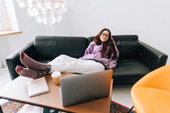 Young caucasian woman relaxing on a sofa in front laptop in living room after work.