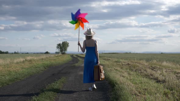 Girl in blue dress with suitcase and pinwheel on country road in summer.